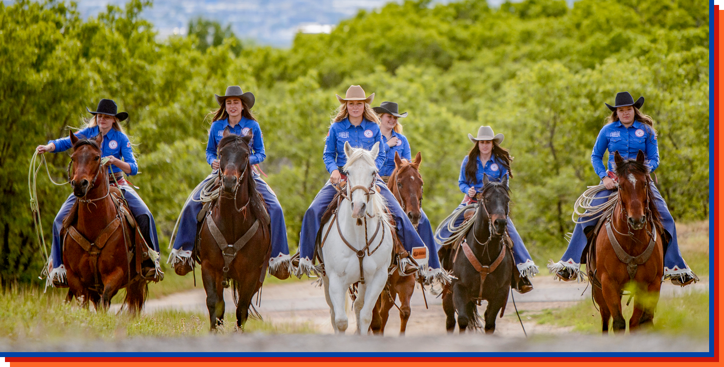 Esta es una foto grupal de las seis vaqueras del Colectivo de Vaqueras de Utah montando a caballo, vistiendo camisas occidentales azules y chaparreras personalizadas en azul, blanco y rojo, con el cielo azul y la hierba verde de fondo.